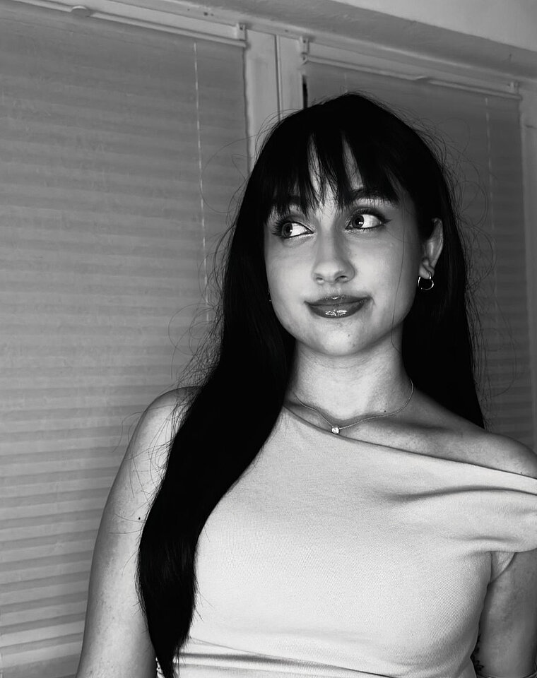 Eine weiblich gelesene Person mit langen dunklen Haaren und Pony blickt zur Seite, im Hintergrund ist eine Jalousie zu sehen